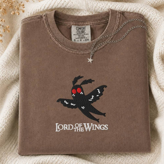 A folded brown sweatshirt with a black moth graphic and the words 'LORD OF THE WINGS' on the front, styled on a cream knit blanket with a silver star pendant necklace.