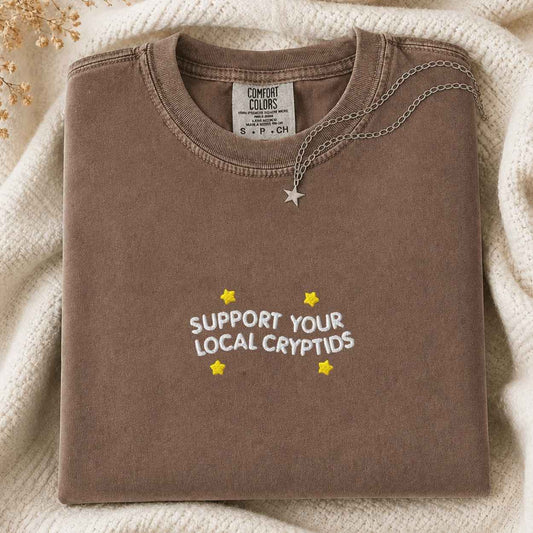 A folded brown sweatshirt with 'SUPPORT YOUR LOCAL CRYPTIDS' and yellow stars printed on the front, styled on a cream knit blanket with a silver star necklace. The neck label shows the brand Comfort Colors.