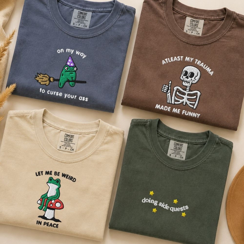 Four folded graphic T-shirts in muted blue, brown, cream, and green, featuring quirky embroidered designs and humorous text.