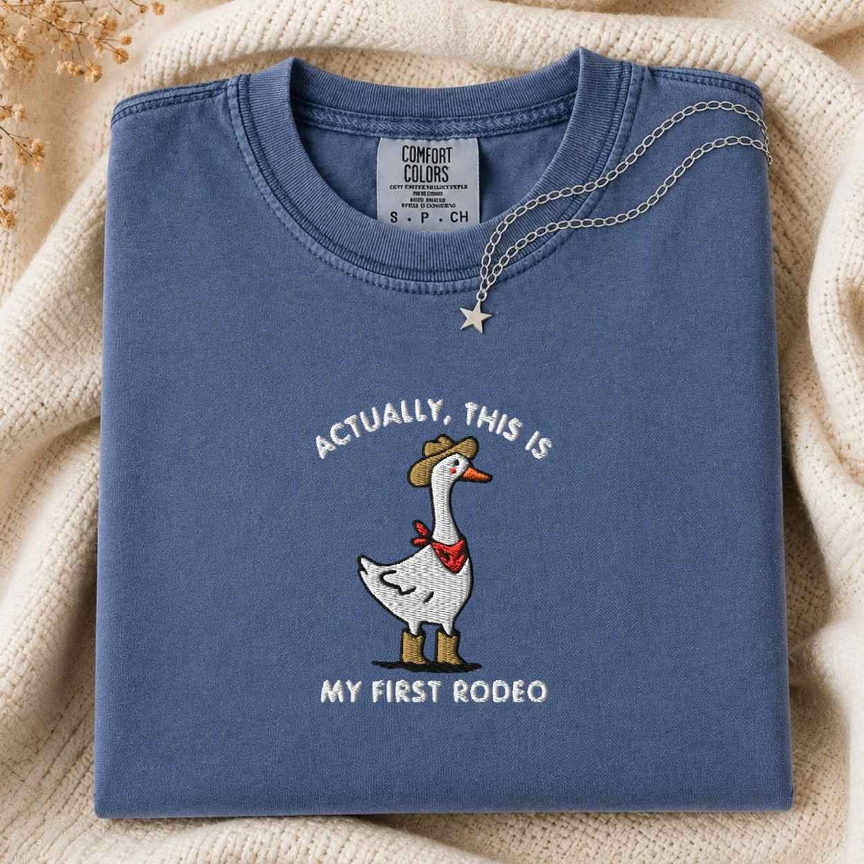 A folded slate blue t-shirt with a western-themed goose graphic and the text 'ACTUALLY, THIS IS MY FIRST RODEO'. The shirt is styled on a cream-colored knit blanket with a silver necklace draped over the collar.