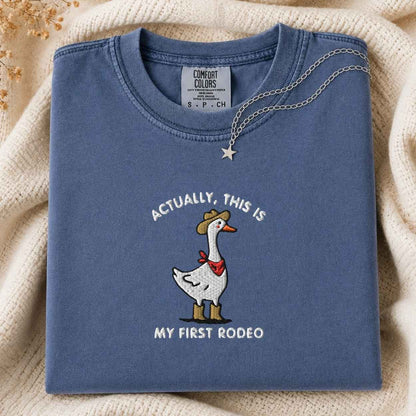 A folded slate blue t-shirt with a western-themed goose graphic and the text 'ACTUALLY, THIS IS MY FIRST RODEO'. The shirt is styled on a cream-colored knit blanket with a silver necklace draped over the collar.