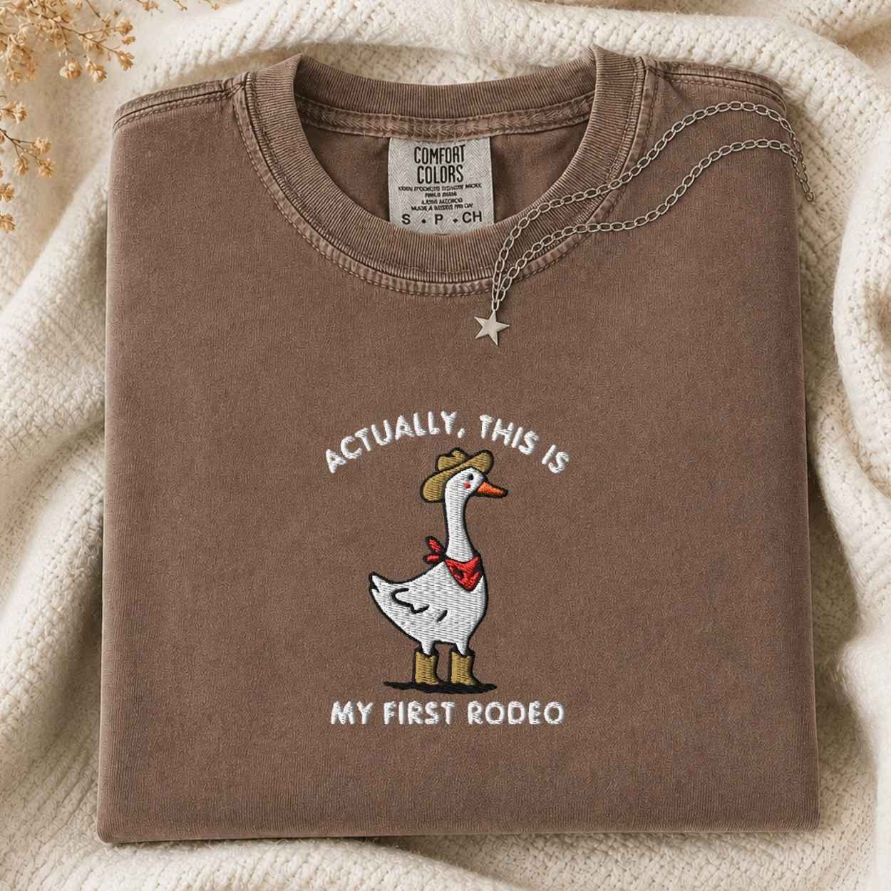 A folded brown t-shirt with a western-themed goose graphic and the text 'ACTUALLY, THIS IS MY FIRST RODEO'. The shirt is styled on a cream-colored knit blanket with a silver necklace draped over the collar.
