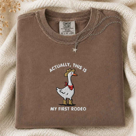 A folded brown t-shirt with a western-themed goose graphic and the text 'ACTUALLY, THIS IS MY FIRST RODEO'. The shirt is styled on a cream-colored knit blanket with a silver necklace draped over the collar.