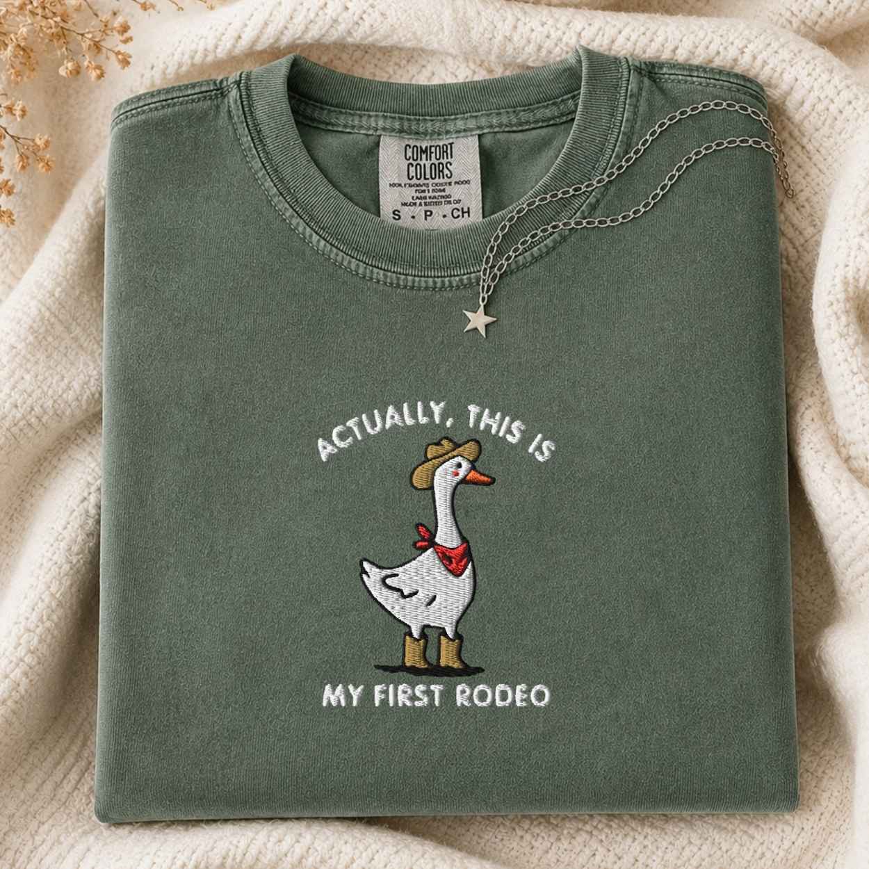 A folded sage green Comfort Colors t-shirt featuring a white goose embroidered with a cowboy hat and red bandana, with the text 'ACTUALLY, THIS IS MY FIRST RODEO'. The shirt is styled on a cream knit blanket with a silver star necklace.