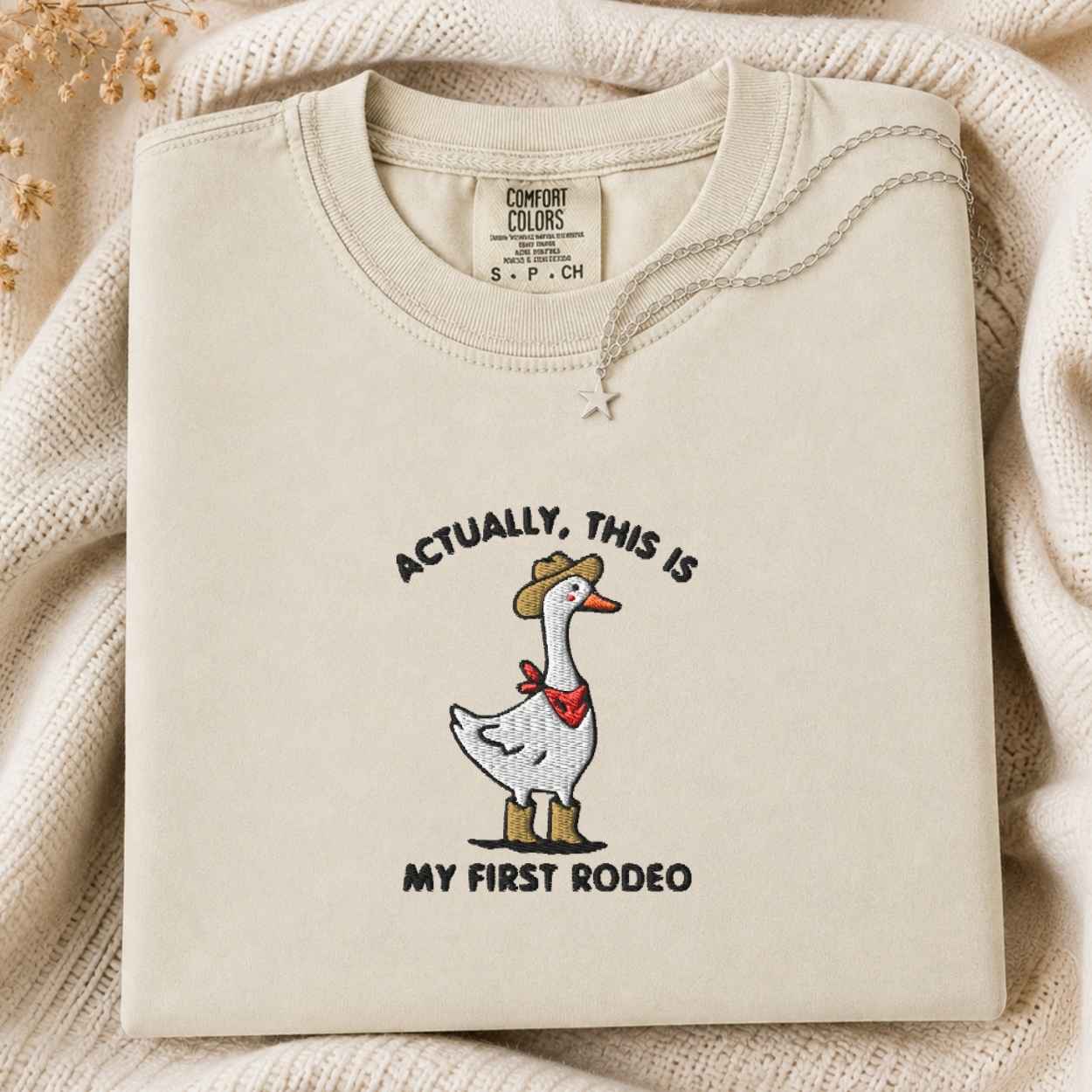 A folded cream tshirt with a graphic of a goose wearing a cowboy hat and red bandana, with the text 'ACTUALLY, THIS IS MY FIRST RODEO'. The sweatshirt is displayed on a cozy cream knit blanket with dried flowers in the corner.