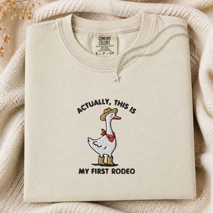 A folded cream tshirt with a graphic of a goose wearing a cowboy hat and red bandana, with the text 'ACTUALLY, THIS IS MY FIRST RODEO'. The sweatshirt is displayed on a cozy cream knit blanket with dried flowers in the corner.
