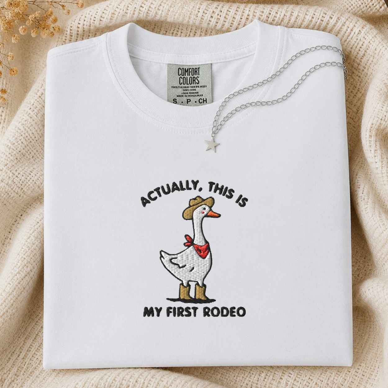 A folded white Comfort Colors t-shirt with a western-themed goose graphic and the text 'ACTUALLY, THIS IS MY FIRST RODEO'. The shirt is styled on a beige knit blanket with a silver necklace draped over the collar.