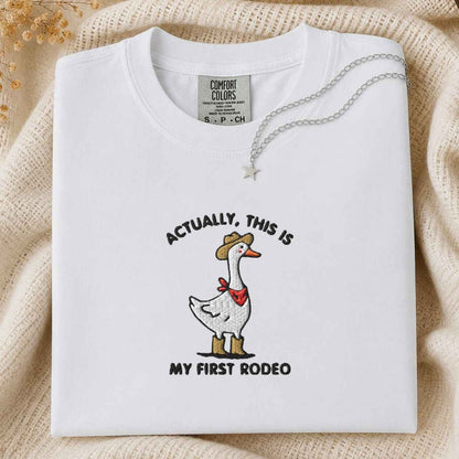 A folded white Comfort Colors t-shirt with a western-themed goose graphic and the text 'ACTUALLY, THIS IS MY FIRST RODEO'. The shirt is styled on a beige knit blanket with a silver necklace draped over the collar.