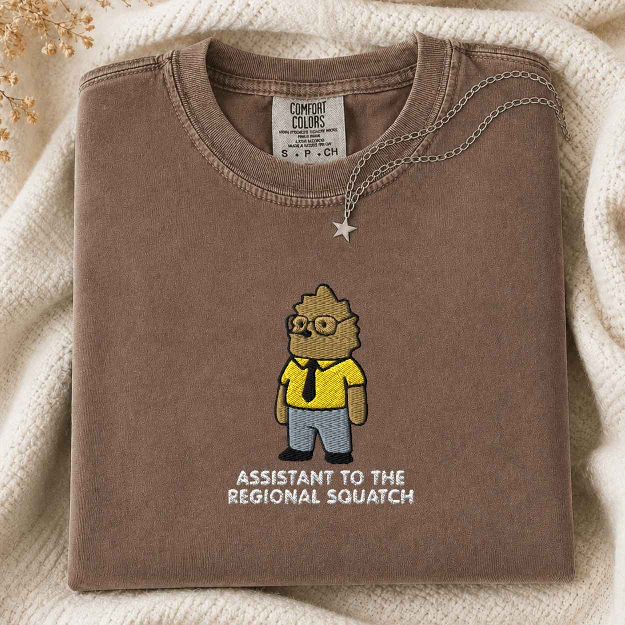 A folded brown sweatshirt with a cartoon graphic of a character in a yellow shirt and blue pants, with the text 'ASSISTANT TO THE REGIONAL SQUATCH' underneath. The sweatshirt is styled on a cream knit blanket with a silver necklace draped over the collar.