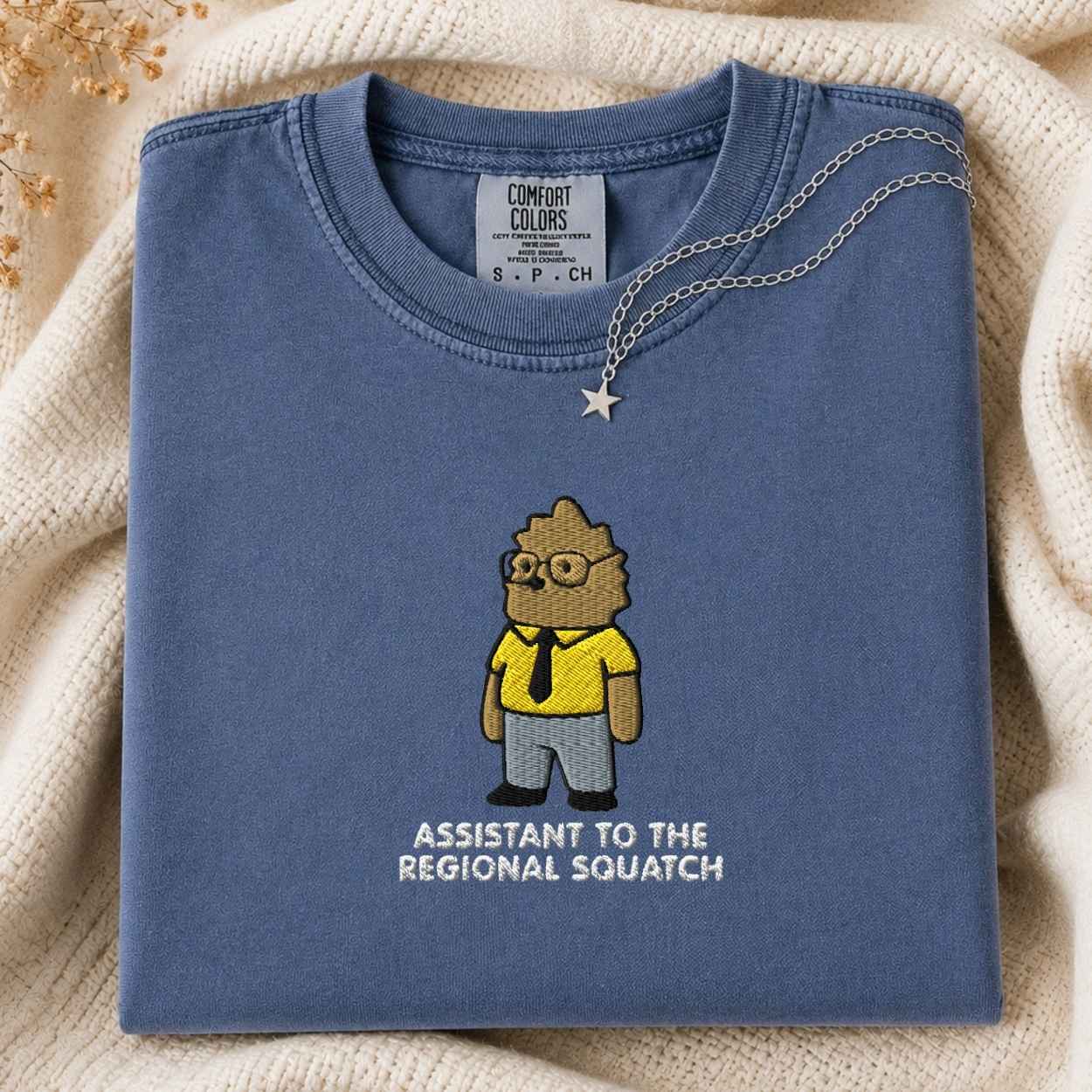A folded slate blue t-shirt with a cartoon graphic of a squatch as an assistant regional squatch, styled on a cream knit blanket with a silver necklace.