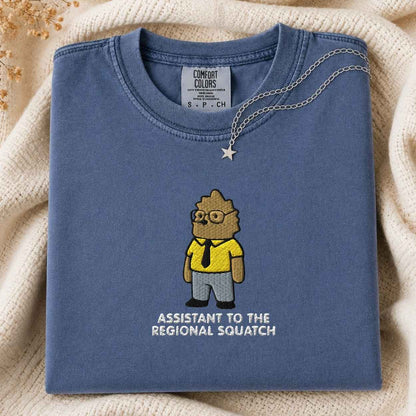 A folded slate blue t-shirt with a cartoon graphic of a squatch as an assistant regional squatch, styled on a cream knit blanket with a silver necklace.
