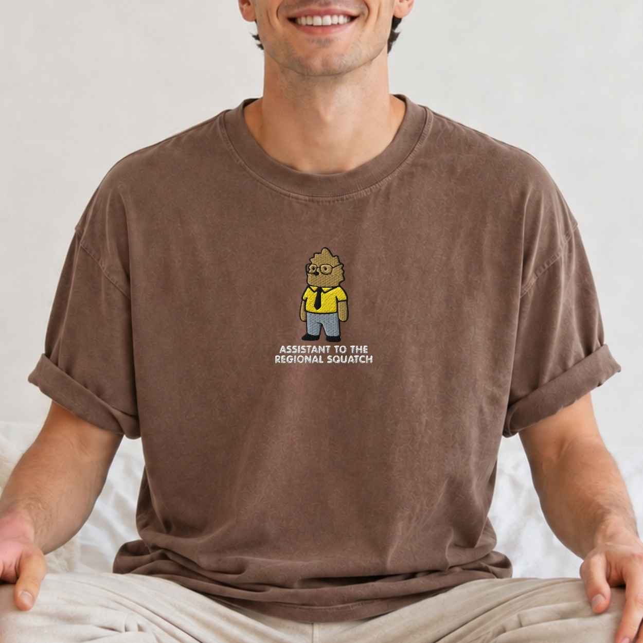 A man wearing a brown crewneck t-shirt with a cartoon graphic of a bigfoot and wearing a straw hat, with the text 'Assistant To The Regional Squatch' printed below. The man is sitting cross-legged against a neutral light background.