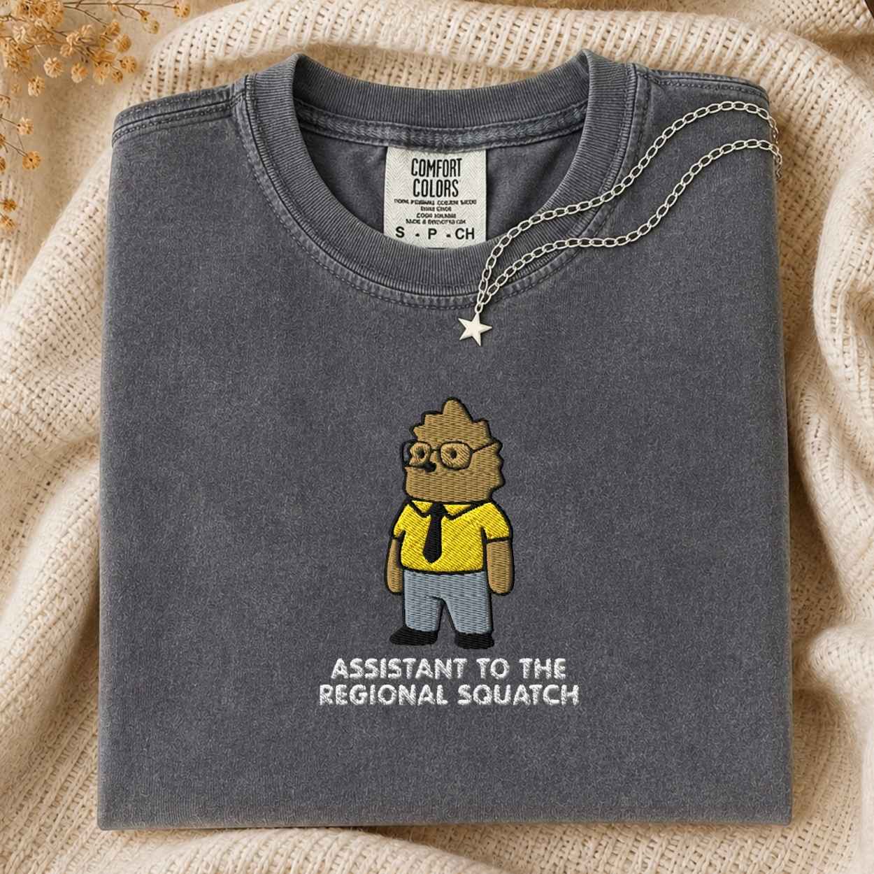A folded dark gray Comfort Colors t-shirt featuring a cartoon graphic of a squatch in a yellow shirt and black tie, with the text 'ASSISTANT TO THE REGIONAL SQUATCH' printed below. A silver necklace with a star pendant is draped over the collar, set against a cozy beige knit background.