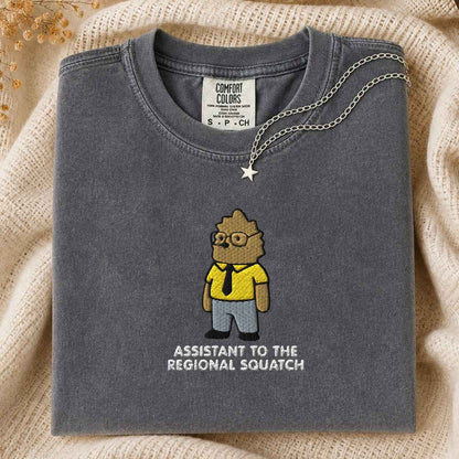 A folded dark gray Comfort Colors t-shirt featuring a cartoon graphic of a squatch in a yellow shirt and black tie, with the text 'ASSISTANT TO THE REGIONAL SQUATCH' printed below. A silver necklace with a star pendant is draped over the collar, set against a cozy beige knit background.