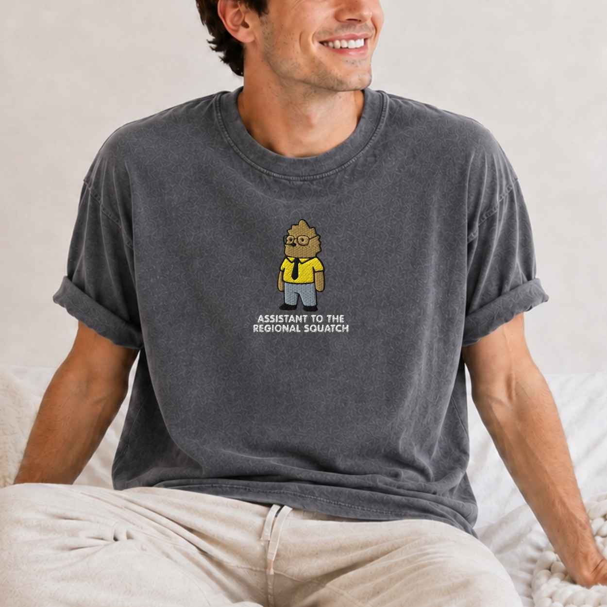 A man wearing a dark gray washed t-shirt with a cartoon bigfoot of a character labeled 'Assistant to the Regional Squatch' sitting on a bed.