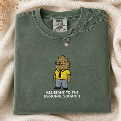 A folded sage green Comfort Colors t-shirt featuring a cartoon graphic of a bigfoot in a yellow shirt and blue pants with the text 'ASSISTANT TO THE REGIONAL SQUATCH' underneath. The shirt is styled on a cream-colored knit blanket with a silver star necklace draped over the collar.