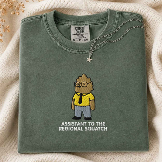 A folded sage green Comfort Colors t-shirt featuring a cartoon graphic of a bigfoot in a yellow shirt and blue pants with the text 'ASSISTANT TO THE REGIONAL SQUATCH' underneath. The shirt is styled on a cream-colored knit blanket with a silver star necklace draped over the collar.