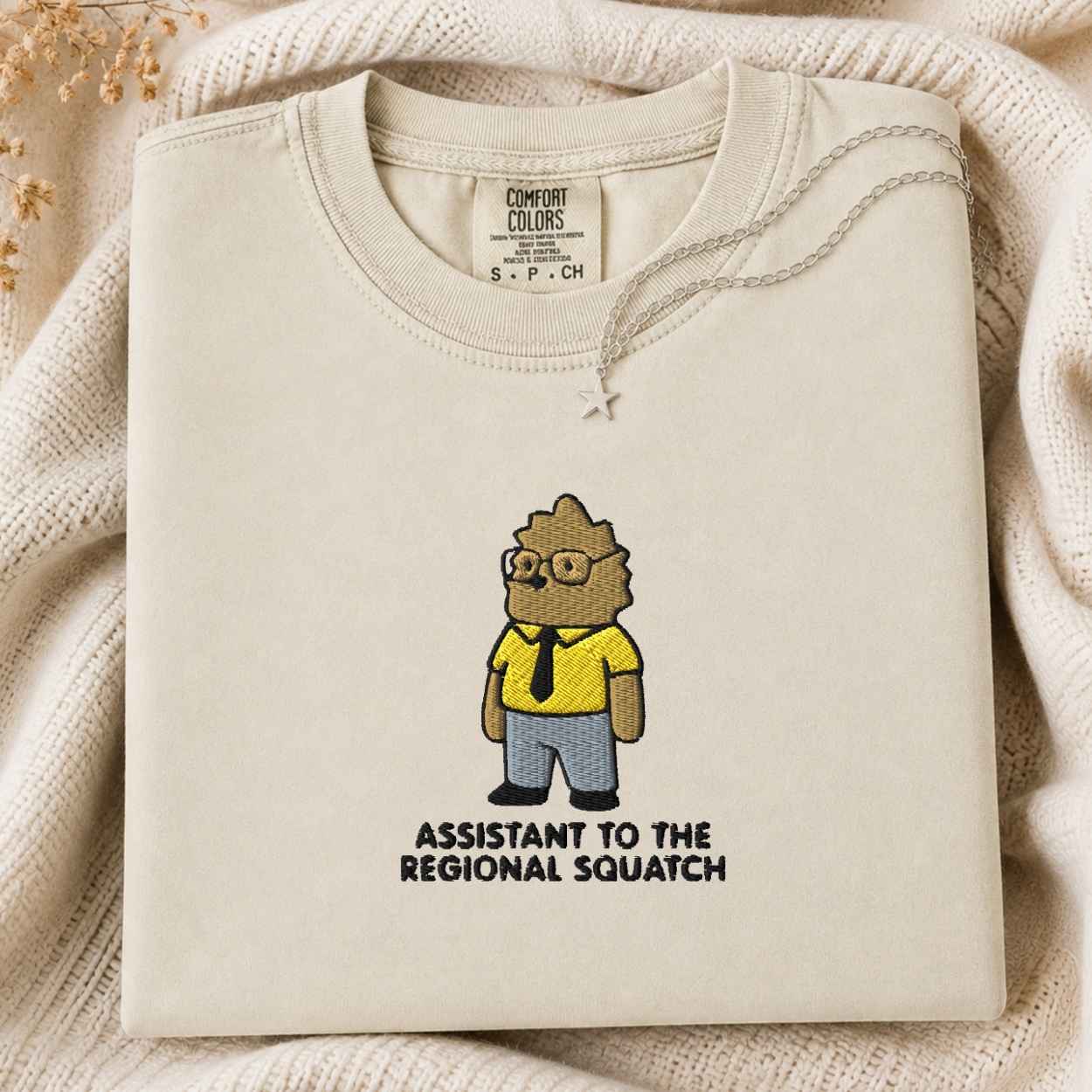 A folded cream-colored tshirt with a cartoon graphic of a squatch wearing glasses and a yellow shirt, with the text 'ASSISTANT TO THE REGIONAL SQUATCH' printed below. The sweatshirt is styled on a cozy cream knit blanket with dried flowers in the corner.
