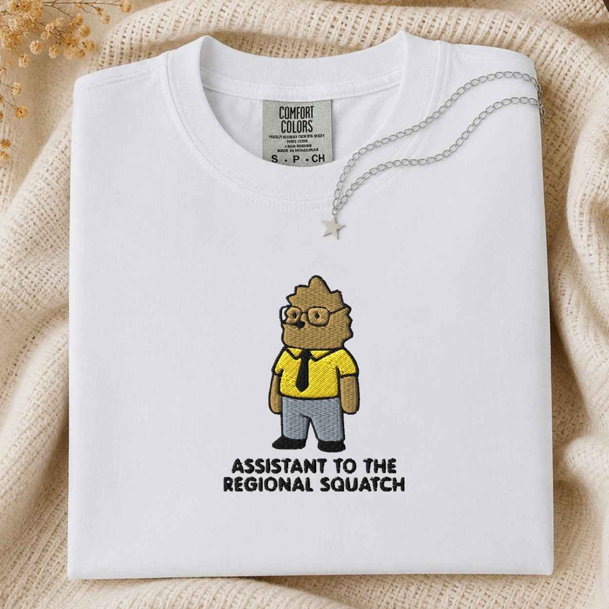 A folded white Comfort Colors t-shirt with a cartoon graphic of a squatch wearing a yellow shirt and gray pants, with the text 'ASSISTANT TO THE REGIONAL SQUATCH' printed below. The shirt is styled on a beige knit blanket with a silver necklace draped over the collar.