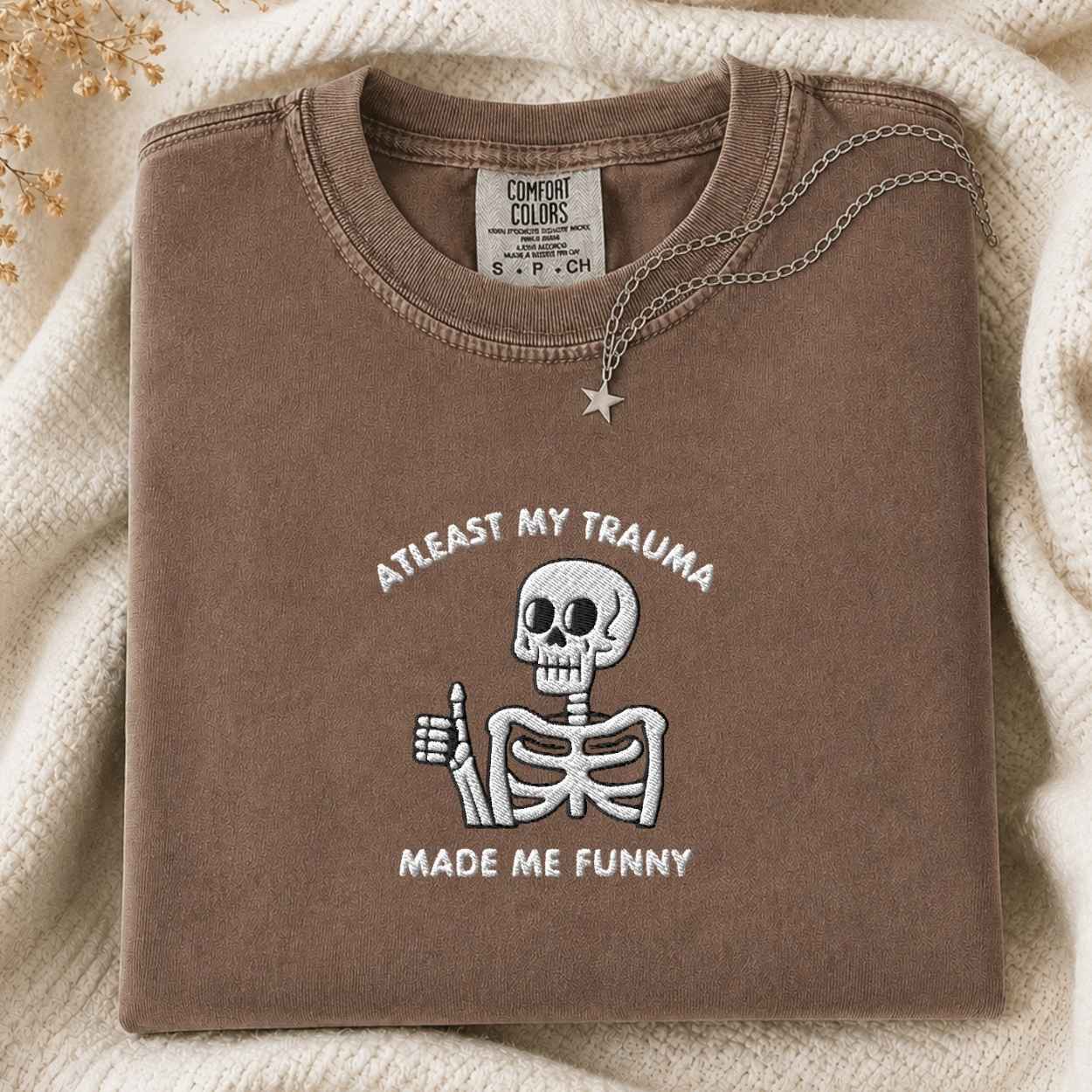 A folded brown crewneck tshirt with a white embroidered cartoon skeleton giving a thumbs-up and the text 'AT LEAST MY TRAUMA MADE ME FUNNY'. The tshirt is styled on a cream knit blanket with a silver star pendant.
