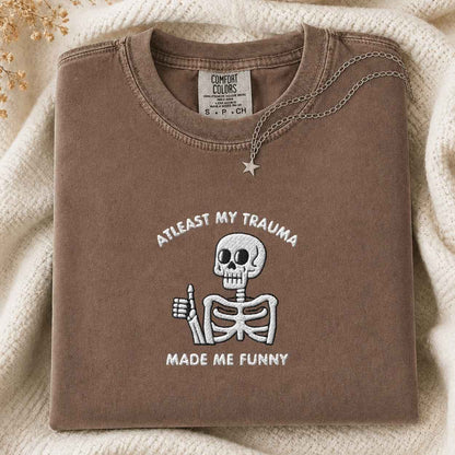 A folded brown crewneck tshirt with a white embroidered cartoon skeleton giving a thumbs-up and the text 'AT LEAST MY TRAUMA MADE ME FUNNY'. The tshirt is styled on a cream knit blanket with a silver star pendant.