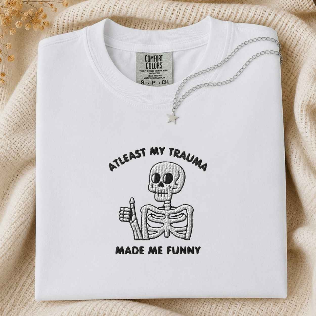 A folded white Comfort Colors t-shirt with a black graphic of a skeleton giving a thumbs-up and the text 'ATLEAST MY TRAUMA MADE ME FUNNY'. The shirt is styled on a beige knit blanket with a silver necklace draped over the collar.