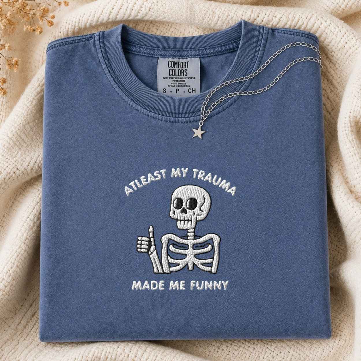 A folded slate blue t-shirt with a white embroidered skeleton giving a thumbs-up and the text 'ATLEAST MY TRAUMA MADE ME FUNNY'. The shirt is styled on a beige knit blanket with a silver necklace draped over the collar.