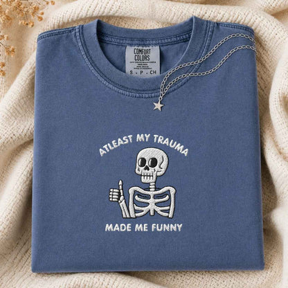 A folded slate blue t-shirt with a white embroidered skeleton giving a thumbs-up and the text 'ATLEAST MY TRAUMA MADE ME FUNNY'. The shirt is styled on a beige knit blanket with a silver necklace draped over the collar.