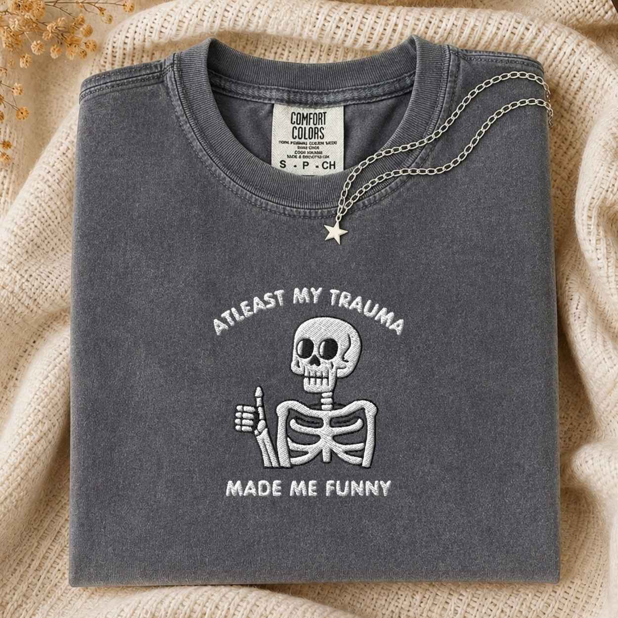 A folded charcoal gray t-shirt with a white embroidered skeleton giving a thumbs-up and the text 'ATLEAST MY TRAUMA MADE ME FUNNY'. A silver star pendant necklace is draped over the collar.