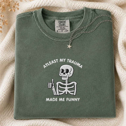 A folded sage green Comfort Colors t-shirt with a white embroidered skeleton giving a thumbs-up and the text 'AT LEAST MY TRAUMA MADE ME FUNNY'. The shirt is styled on a cream knit blanket with a silver star necklace draped over the collar.