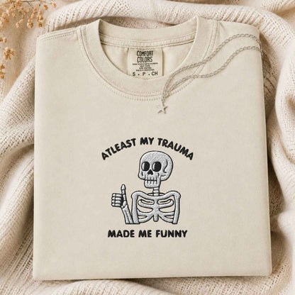 A folded cream-colored crewneck tshirt with a black graphic of a skeleton giving a thumbs-up and the text 'AT LEAST MY TRAUMA MADE ME FUNNY'. The tshirt is displayed on a cozy, textured cream knit blanket with dried flowers in the corner.