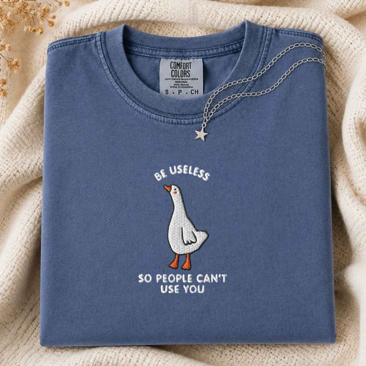 A folded slate blue crewneck t-shirt with a white goose graphic and the text 'BE USELESS SO PEOPLE CAN'T USE YOU' in white. A silver star necklace is draped over the collar.