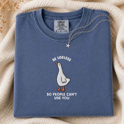 A folded slate blue crewneck t-shirt with a white goose graphic and the text 'BE USELESS SO PEOPLE CAN'T USE YOU' in white. A silver star necklace is draped over the collar.