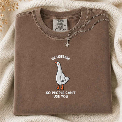 A folded brown t-shirt with a white goose graphic and the text 'BE USELESS SO PEOPLE CAN'T USE YOU' in white, styled on a cream knit blanket with a silver star necklace.