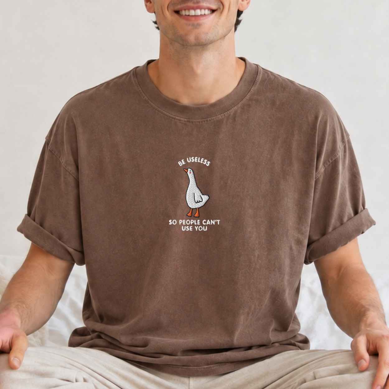 A man wearing a brown crewneck t-shirt with a cartoon graphic of a goose, with the text 'Be Useless So People Can't Use You' printed below. The man is sitting cross-legged against a neutral light background.
