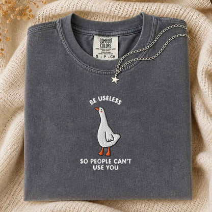 A folded dark gray t-shirt with a white goose graphic and the text 'BE USELESS SO PEOPLE CAN'T USE YOU' in white and orange. A silver chain necklace with a star pendant is draped over the collar.