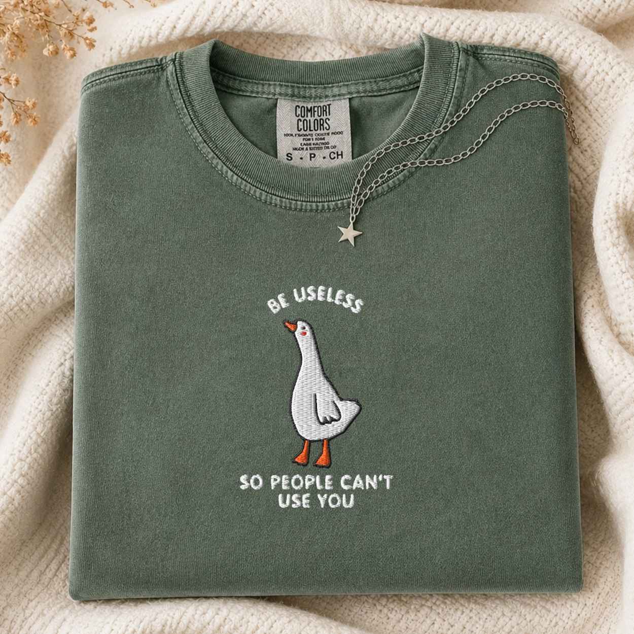 A folded sage green t-shirt with a graphic of a white goose and the text 'BE USELESS SO PEOPLE CAN'T USE YOU' in white and orange. The shirt is styled on a cream-colored knit blanket with a silver star necklace draped over the collar.