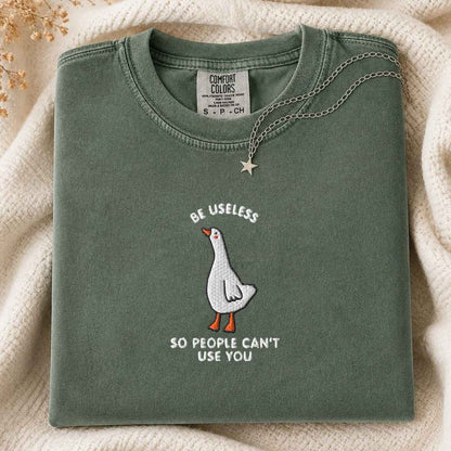 A folded sage green t-shirt with a graphic of a white goose and the text 'BE USELESS SO PEOPLE CAN'T USE YOU' in white and orange. The shirt is styled on a cream-colored knit blanket with a silver star necklace draped over the collar.