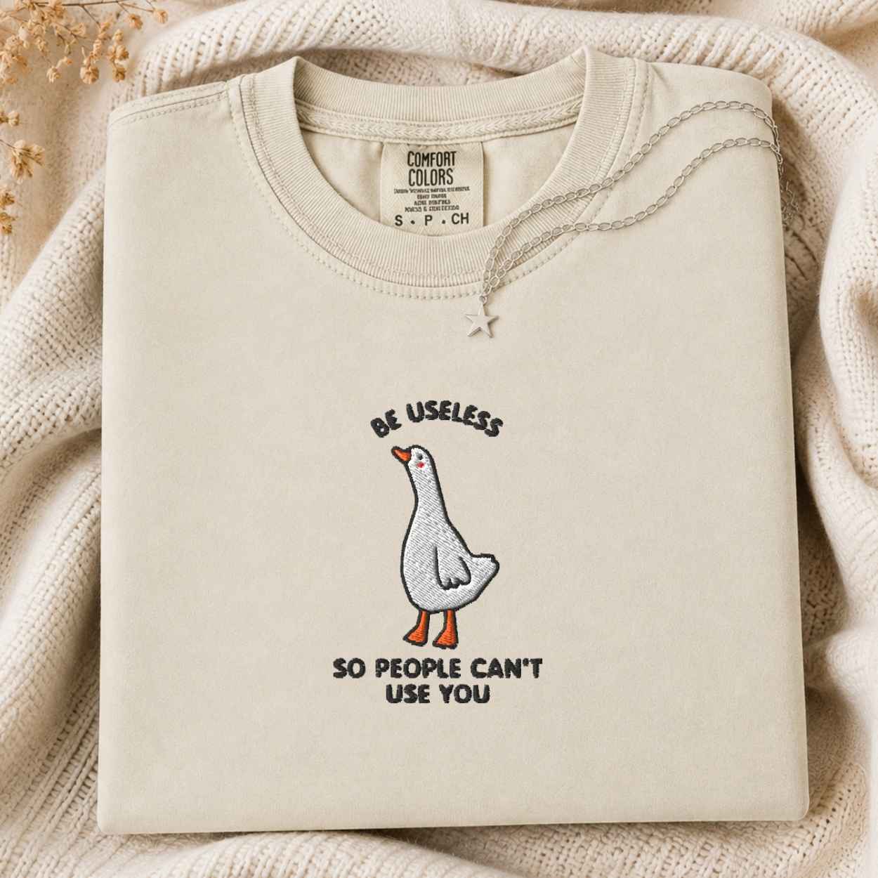 A folded cream-colored tshirt with a graphic of a goose and the text 'BE USELESS SO PEOPLE CAN'T USE YOU' on a cozy knit blanket background.