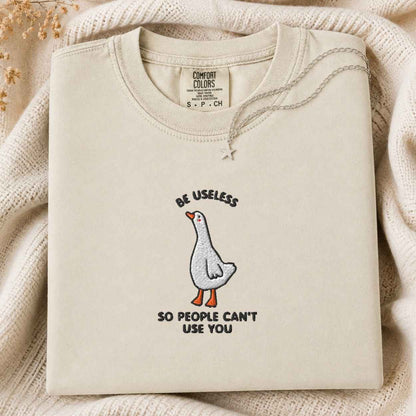 A folded cream-colored tshirt with a graphic of a goose and the text 'BE USELESS SO PEOPLE CAN'T USE YOU' on a cozy knit blanket background.