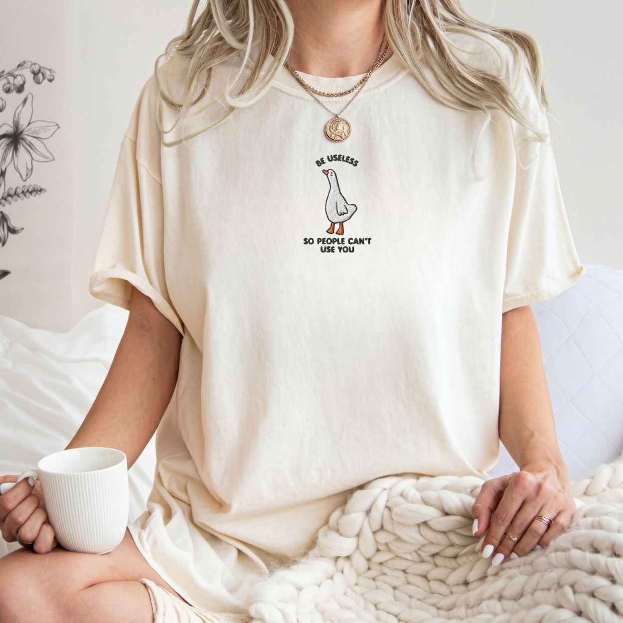 A woman wearing a cream-colored oversized t-shirt with a small graphic of a goose and the text 'BE USELESS SO I CAN'T USE YOU' on the front, sitting in a cozy indoor setting and holding a white mug.