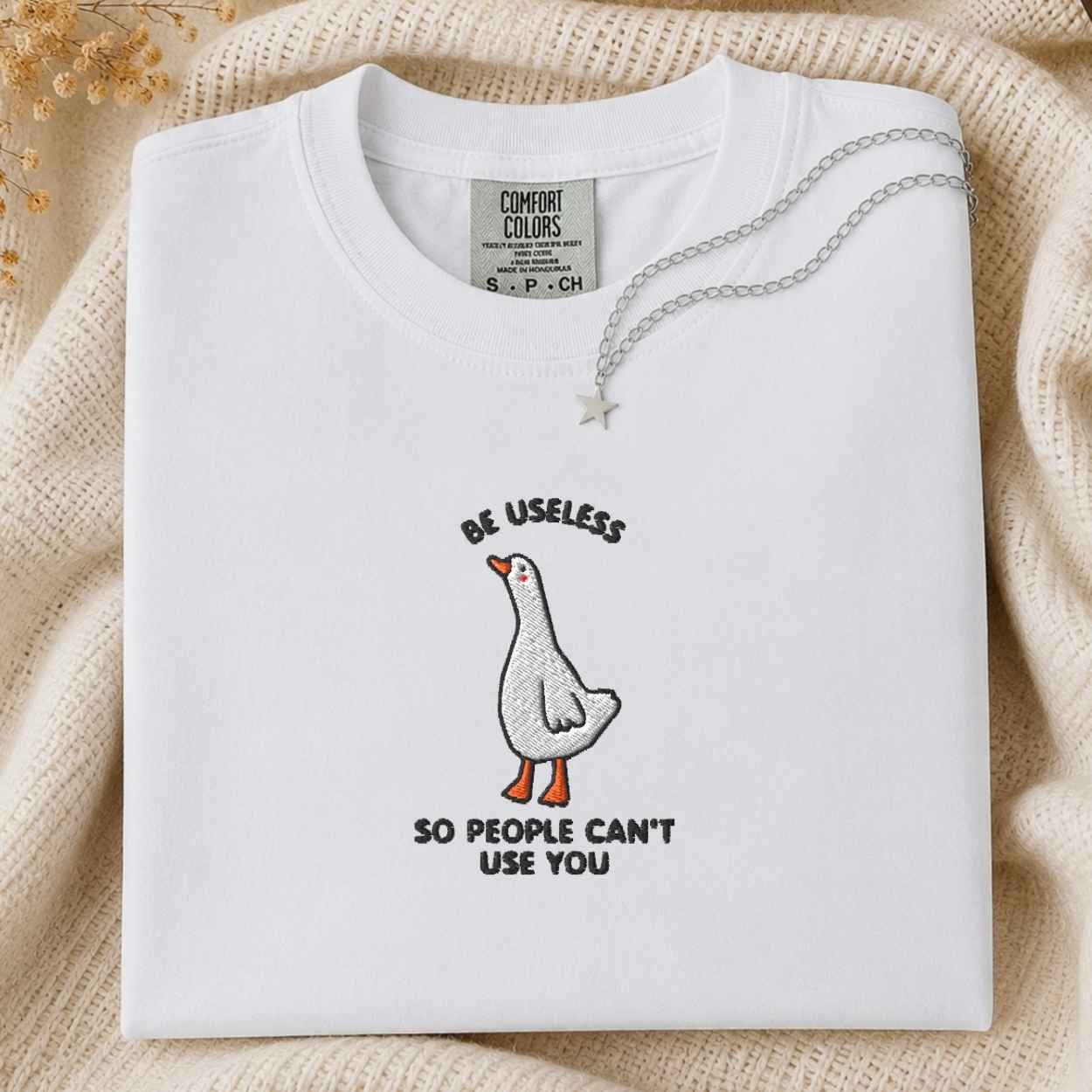 A folded white Comfort Colors t-shirt with a humorous goose graphic and the text 'BE USELESS SO PEOPLE CAN'T USE YOU'. The shirt is styled on a beige knit blanket with a silver necklace draped over the collar.