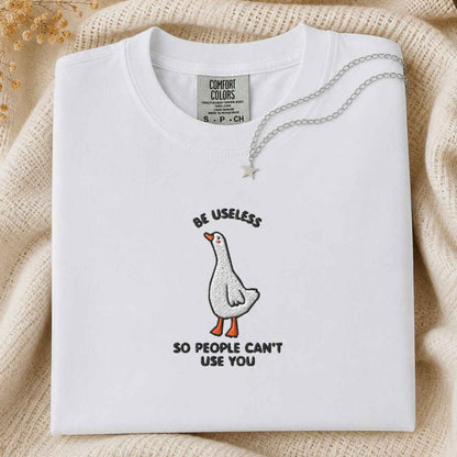 A folded white Comfort Colors t-shirt with a humorous goose graphic and the text 'BE USELESS SO PEOPLE CAN'T USE YOU'. The shirt is styled on a beige knit blanket with a silver necklace draped over the collar.