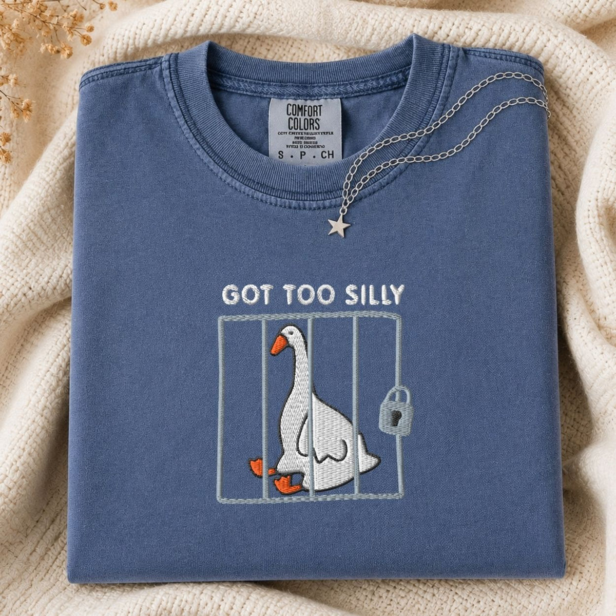 A folded slate blue t-shirt with a graphic of a silly goose in a cage and the text 'GOT TOO SILLY'. A silver necklace with a star pendant is draped over the collar.