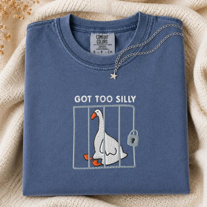 A folded slate blue t-shirt with a graphic of a silly goose in a cage and the text 'GOT TOO SILLY'. A silver necklace with a star pendant is draped over the collar.