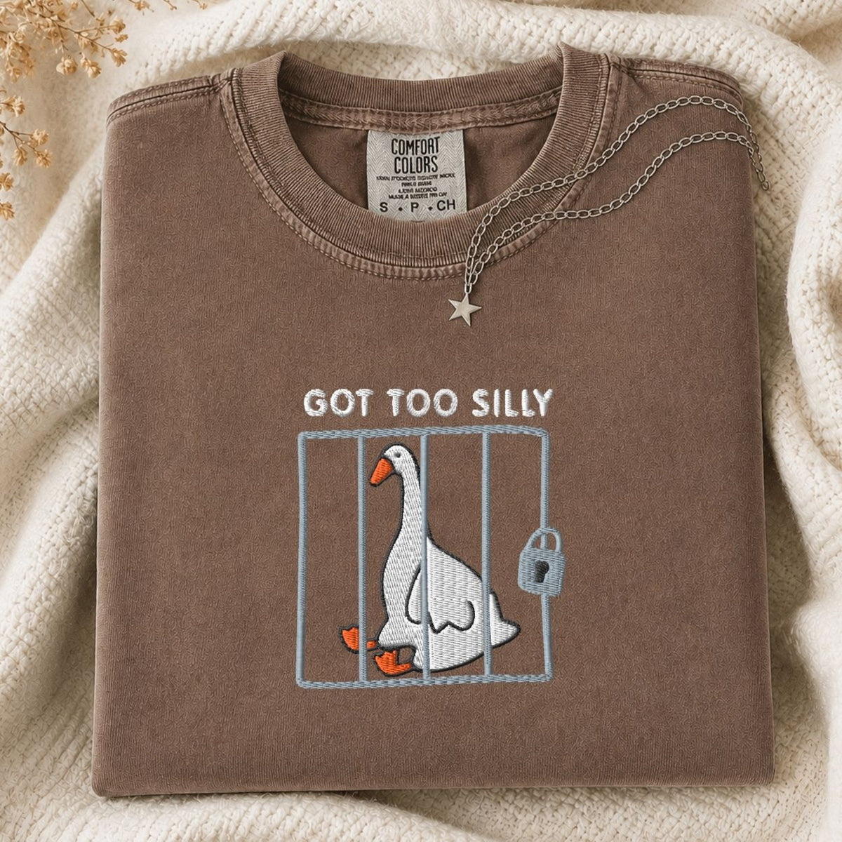 A folded brown tshirt with a graphic of a silly goose in a cage and the text 'GOT TOO SILLY' on the front, styled on a cream knit blanket with a necklace draped over the collar.
