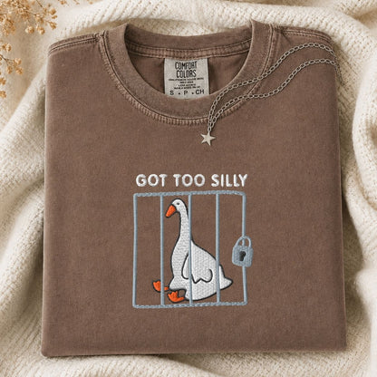 A folded brown tshirt with a graphic of a silly goose in a cage and the text 'GOT TOO SILLY' on the front, styled on a cream knit blanket with a necklace draped over the collar.