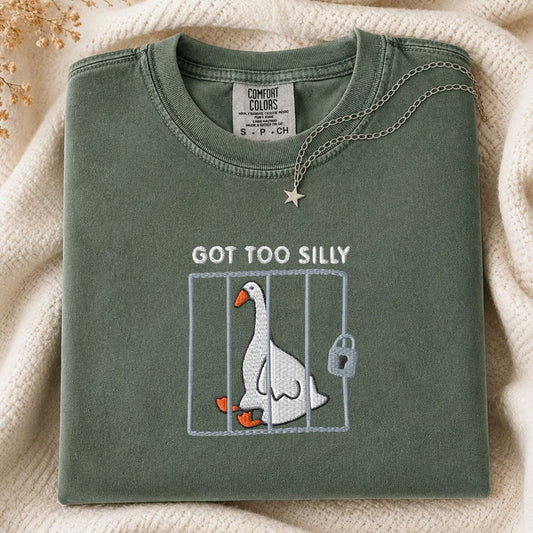 A folded sage green Comfort Colors t-shirt with a graphic of a goose in a cage and the text 'GOT TOO SILLY'. The shirt is styled on a cream-colored knit blanket with a gold star necklace draped over the collar.