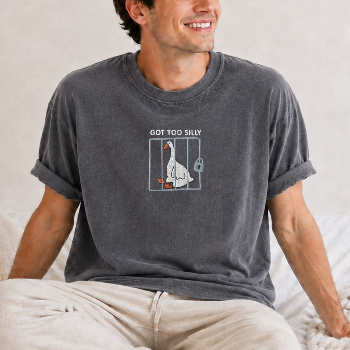 A man wearing a dark gray washed t-shirt with the design of a goose in a cage and the text 'Got Too Silly' printed on the chest, sitting on a bed.