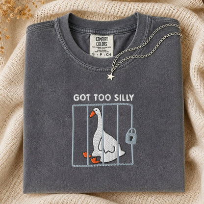 A folded dark gray t-shirt with a graphic of a white silly goose in a cage and the text 'GOT TOO SILLY' on the front, styled on a beige knit blanket with a silver necklace.
